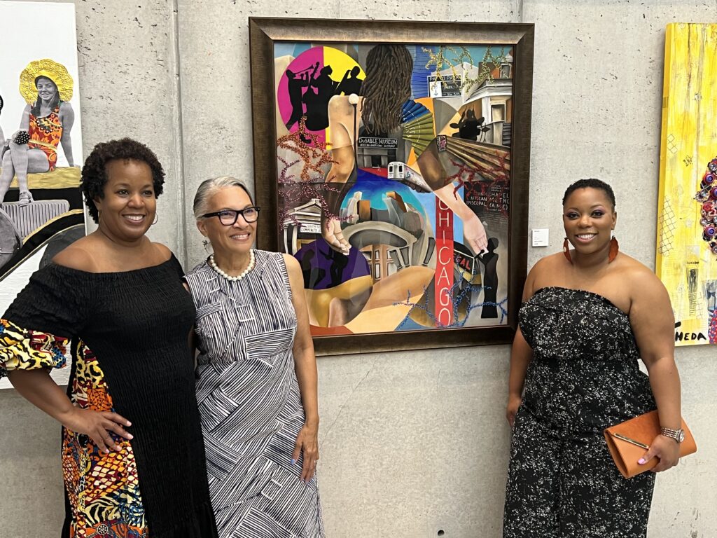 Three women pose, smiling in front of a colorful framed art piece at Nimbus Arts Center